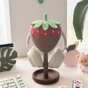 Pu&ograve; includere: Un supporto per cuffie a forma di fragola, rosso, marrone e verde, che tiene delle cuffie bianche. Il supporto ha una base rotonda e uno stelo. Il design ricorda una fragola ricoperta di cioccolato. Su una scrivania con tastiera e accessori.