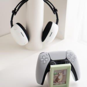May include: A white and black headset rests on a white cylindrical stand. A gray and black game controller is in the foreground, with a small green picture frame holding a cat photo.