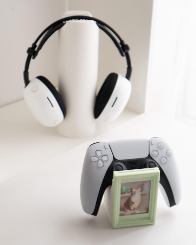 May include: A white and black headset rests on a white cylindrical stand. A gray and black game controller is in the foreground, with a small green picture frame holding a cat photo.
