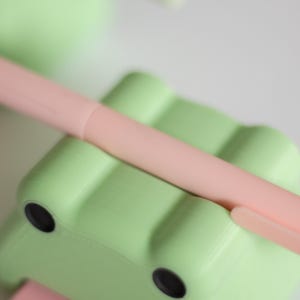 May include: A light green frog-shaped pencil holder with two black eyes. A pink pen is resting on top of the holder.