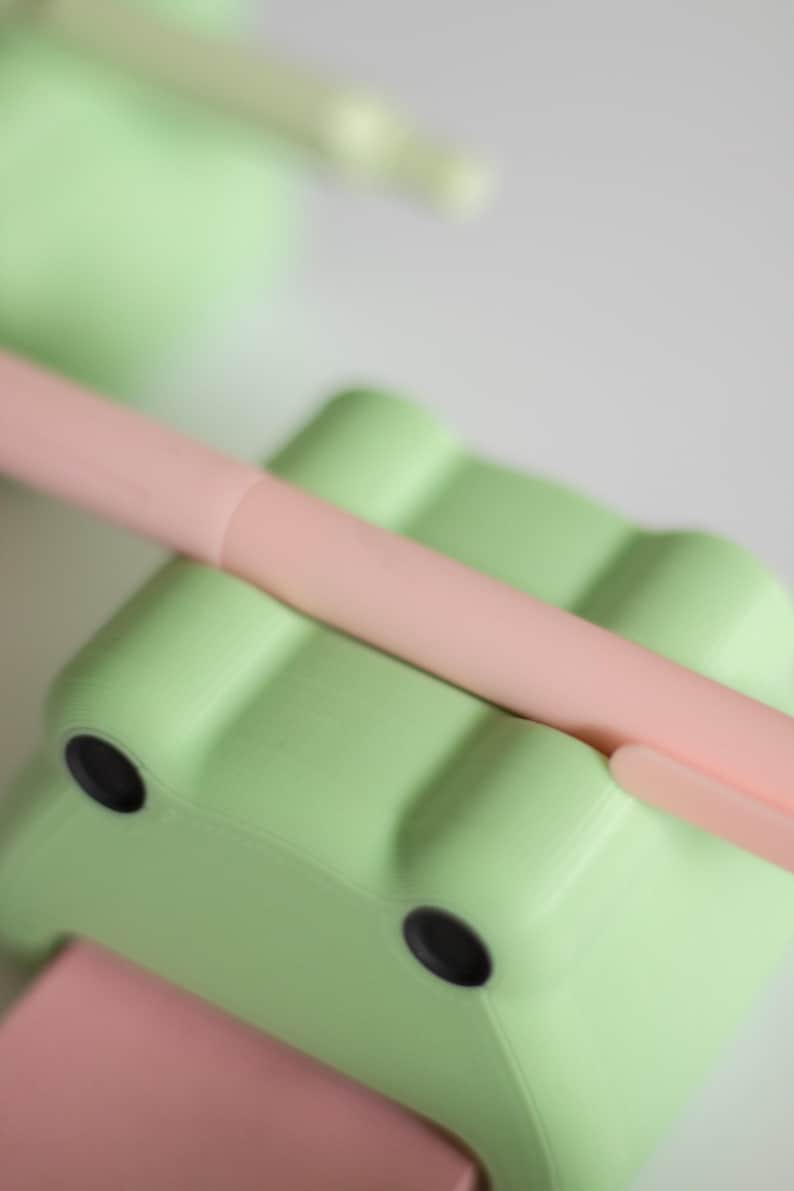 May include: A light green frog-shaped pencil holder with two black eyes. A pink pen is resting on top of the holder.