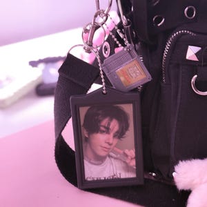 May include: Black backpack with silver hardware and a black strap. Attached are a photo keychain and a small video game cartridge keychain. The photo keychain features a picture of a person.
