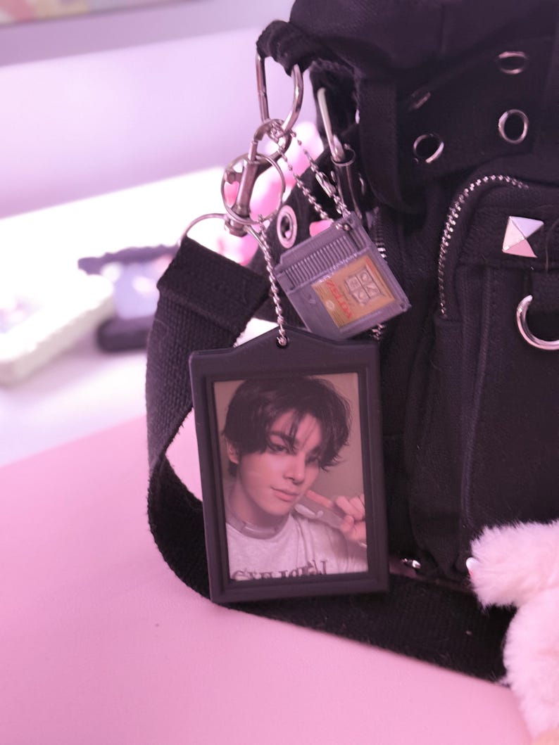 May include: Black backpack with silver hardware and a black strap. Attached are a photo keychain and a small video game cartridge keychain. The photo keychain features a picture of a person.