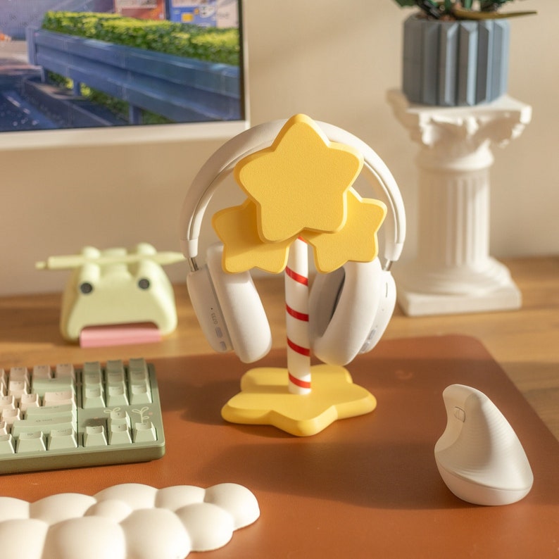 May include: A yellow star-shaped headphone stand with a red and white striped pole, holding white headphones. The stand is on a brown desk with a keyboard, mouse, and other desk accessories.