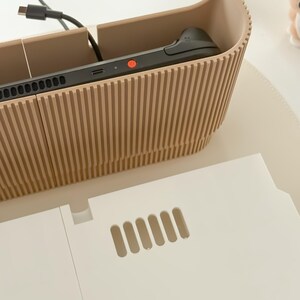 May include: A beige, ribbed-textured organizer with a gaming device inside. A white, rectangular piece with ventilation slots is in front of the organizer. A black cable is connected to the device.