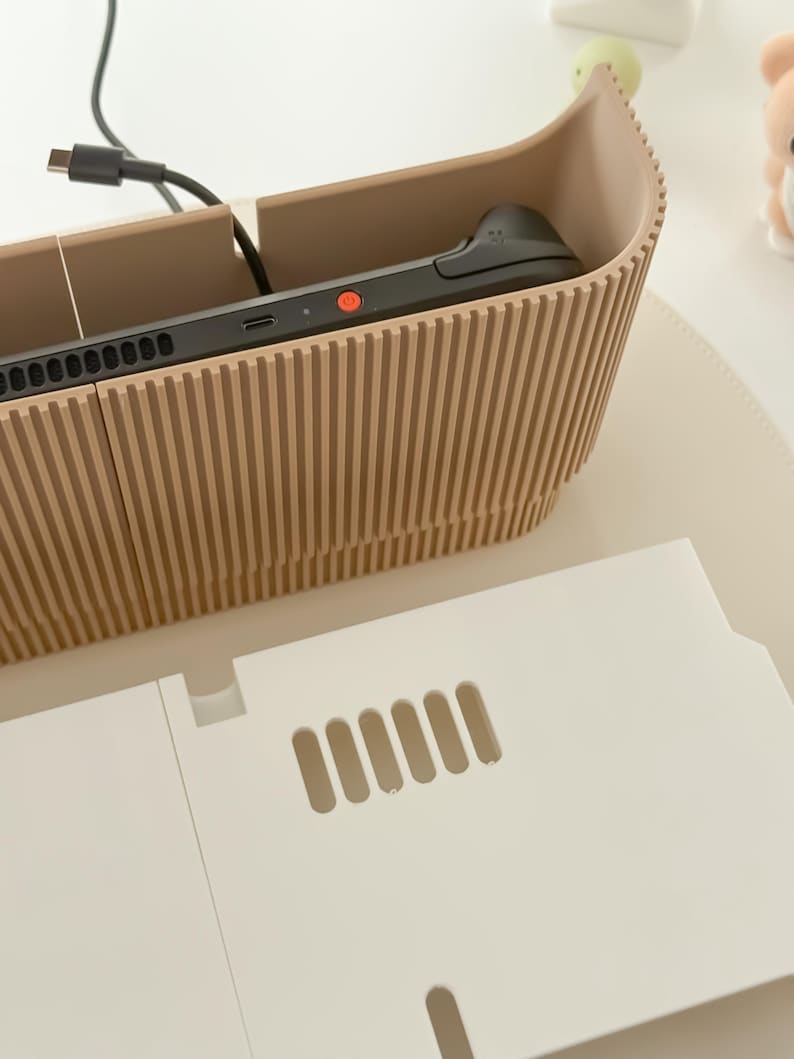 May include: A beige, ribbed-textured organizer with a gaming device inside. A white, rectangular piece with ventilation slots is in front of the organizer. A black cable is connected to the device.