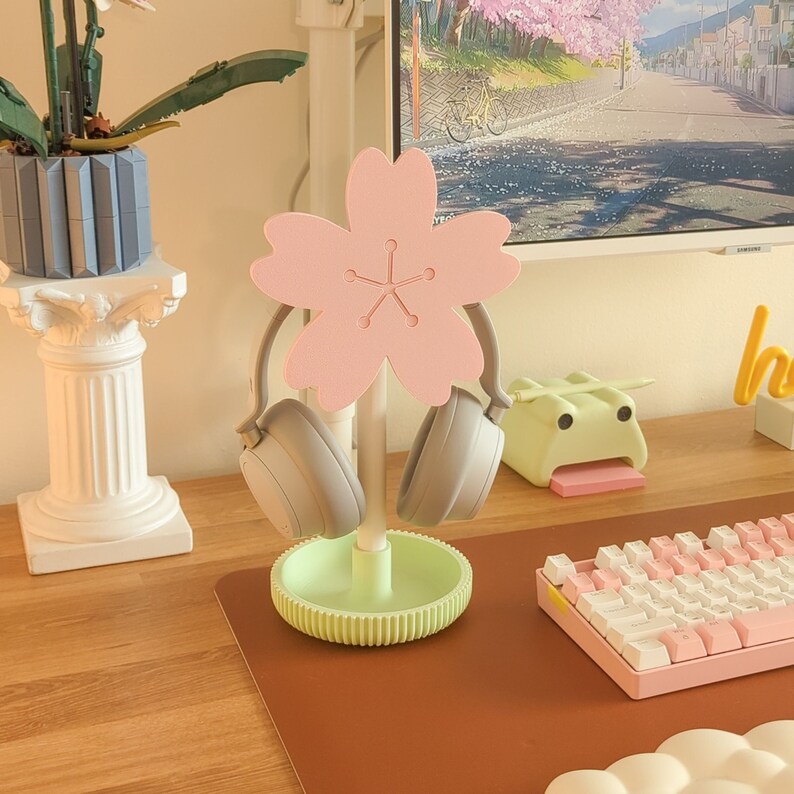 May include: A pink cherry blossom-shaped headphone stand with two gray headphones on it. The stand has a green base and is on a brown surface.
