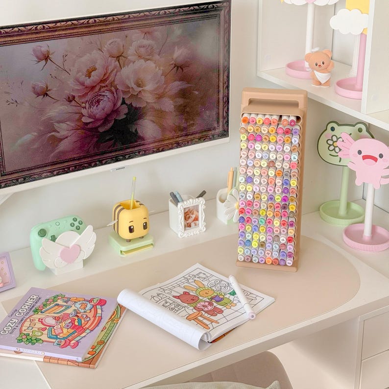 May include: A desk setup featuring a variety of art supplies and decorative items. A tall, beige marker holder is filled with colorful markers. A coloring book and a framed floral print are also visible, along with other cute desk accessories.