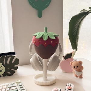Pu&ograve; includere: Un supporto per cuffie bianco a forma di fragola, che tiene cuffie bianche. La fragola &egrave; rossa con un rivestimento marrone simile al cioccolato e foglie verdi. Altri oggetti includono una tastiera, controller di gioco e oggetti decorativi.
