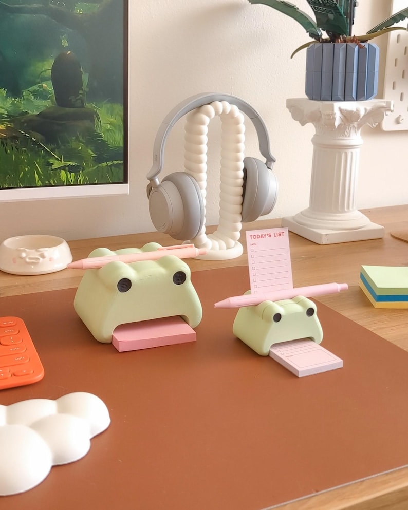 May include: Two green frog-shaped desk organizers with pink sticky notes and a pen on a brown desk mat. The organizers are used to hold notes and pens.