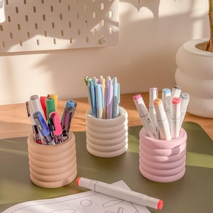 May include: Three ribbed ceramic pen holders in beige, white, and pink, each filled with pens and markers. A white and red marker lies on a coloring page with a cartoon cat illustration. A pegboard and plant are in the background.