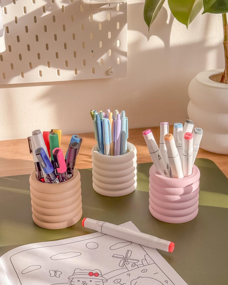 May include: Three ribbed ceramic pen holders in beige, white, and pink, each filled with pens and markers. A white and red marker lies on a coloring page with a cartoon cat illustration. A pegboard and plant are in the background.