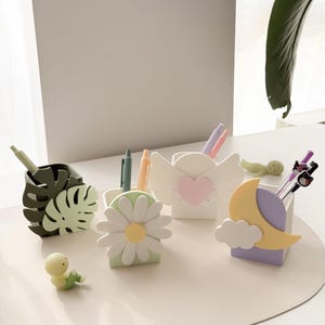 May include: A collection of decorative pen holders in various shapes, including a monstera leaf, daisy, angel wings with a heart, and a moon with a cloud. Each holder contains pens and pencils. A small, yellow figurine is also present.