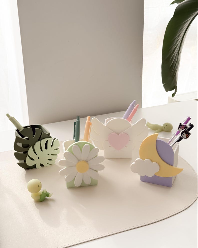 May include: A collection of decorative pen holders in various shapes, including a monstera leaf, daisy, angel wings with a heart, and a moon with a cloud. Each holder contains pens and pencils. A small, yellow figurine is also present.