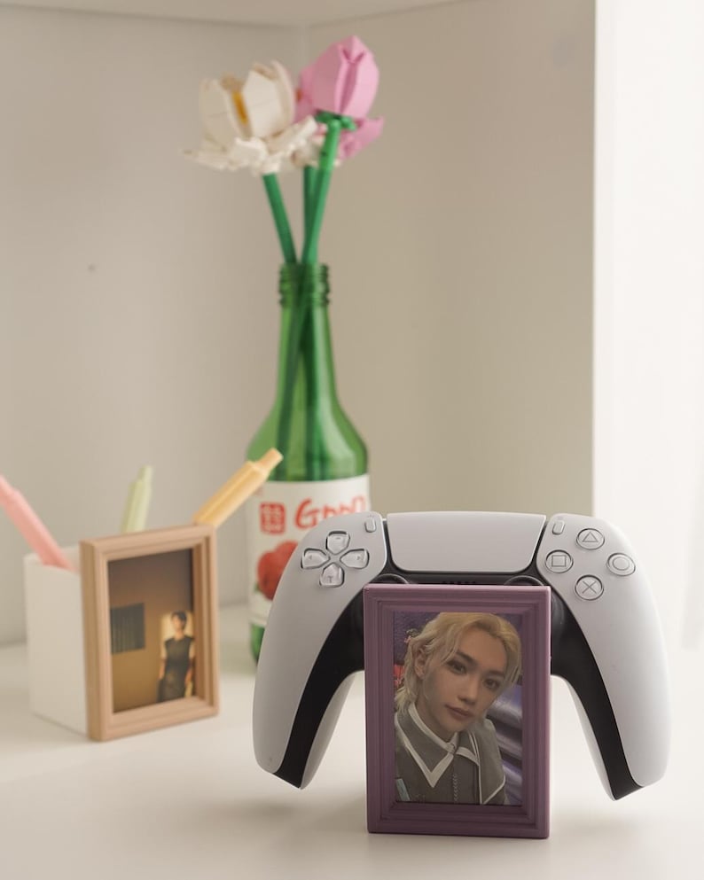 May include: A white and black PlayStation 5 controller with a purple framed photo in front. A green bottle holds Lego flowers. A white container holds pens and a framed photo.
