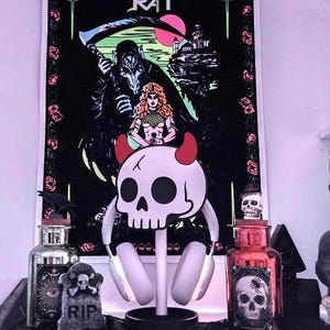 May include: A decorative display featuring a black poster with a gothic illustration of a grim reaper, a woman, and a castle, with the text "CASTLE RAT". A skull-shaped headphone stand with red horns and a pair of headphones is in front of the poster.