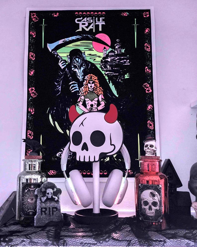 May include: A decorative display featuring a black poster with a gothic illustration of a grim reaper, a woman, and a castle, with the text "CASTLE RAT". A skull-shaped headphone stand with red horns and a pair of headphones is in front of the poster.