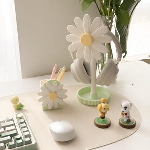 Pu&ograve; includere: Un supporto per cuffie a forma di margherita bianca con centro giallo sostiene un paio di cuffie grigie. Sono presenti anche un portapenne abbinato e accessori da scrivania, tra cui una tastiera, un mouse e delle statuette.