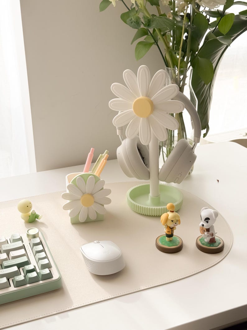 Pu&ograve; includere: Un supporto per cuffie a forma di margherita bianca con centro giallo sostiene un paio di cuffie grigie. Sono presenti anche un portapenne abbinato e accessori da scrivania, tra cui una tastiera, un mouse e delle statuette.