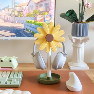 May include: A yellow sunflower-shaped headphone stand with a green base. Two gray headphones are hanging on the stand.