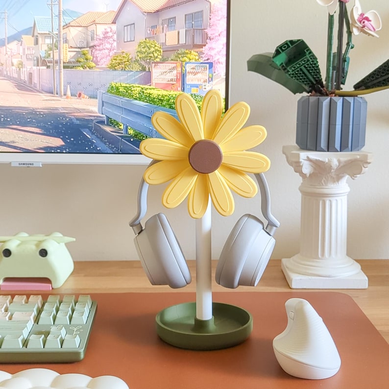 May include: A yellow sunflower-shaped headphone stand with a green base. Two gray headphones are hanging on the stand.