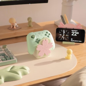 Op de afbeelding: Een lichtgroene gamecontroller rust op een standaard met een roze kersenbloesemontwerp. In de buurt toont een smartphone de tijd en het weer op een roze axolotl-vormige standaard, vergezeld door kleine figuurtjes op een bureau.