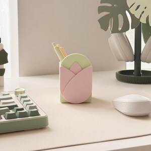 May include: A desk organizer shaped like a tulip, in pastel pink and green, holds pens. A matching keyboard and mouse sit on a light beige desk mat. A decorative plant stand with headphones completes the setup, creating a stylish workspace.