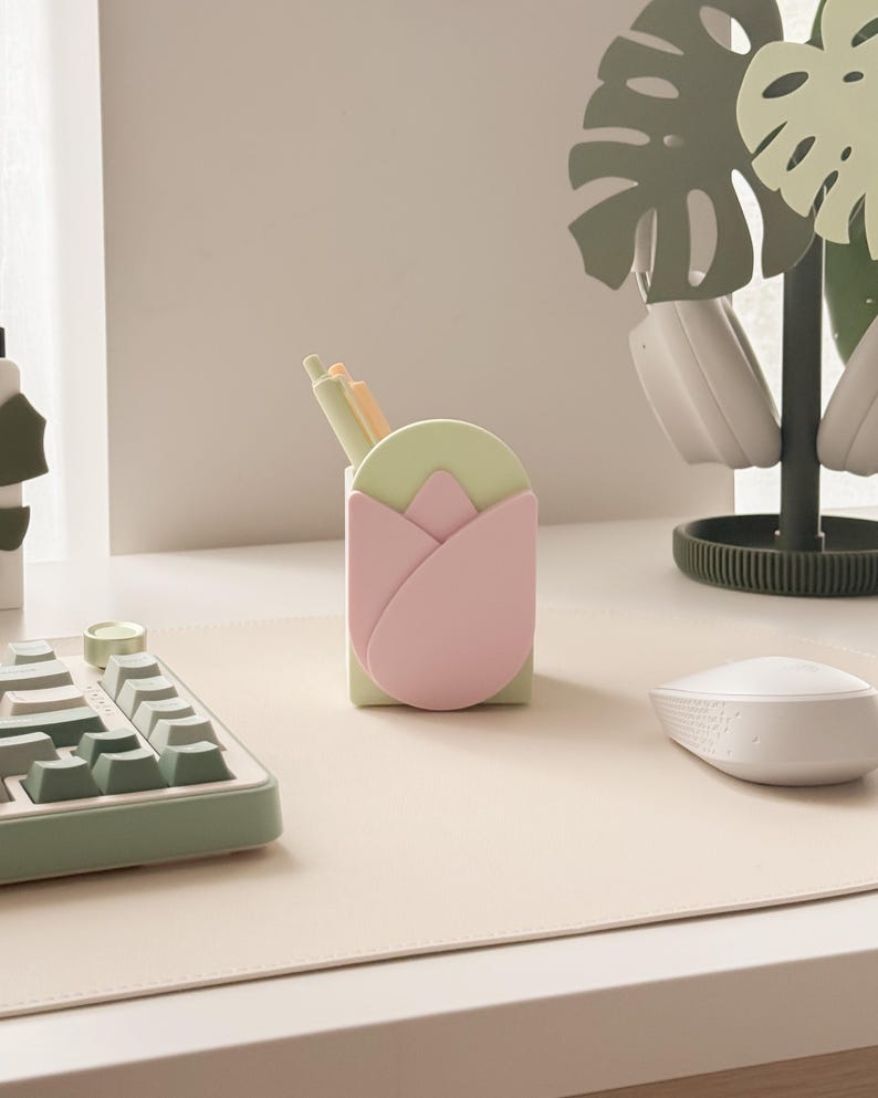 May include: A desk organizer shaped like a tulip, in pastel pink and green, holds pens. A matching keyboard and mouse sit on a light beige desk mat. A decorative plant stand with headphones completes the setup, creating a stylish workspace.