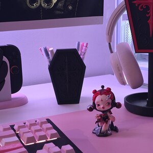 May include: A black coffin-shaped pen holder with a decorative cross design, filled with pens and markers, sits on a pink desk. A small figurine with red hair and a keyboard are also on the desk. A pair of white headphones hangs nearby.