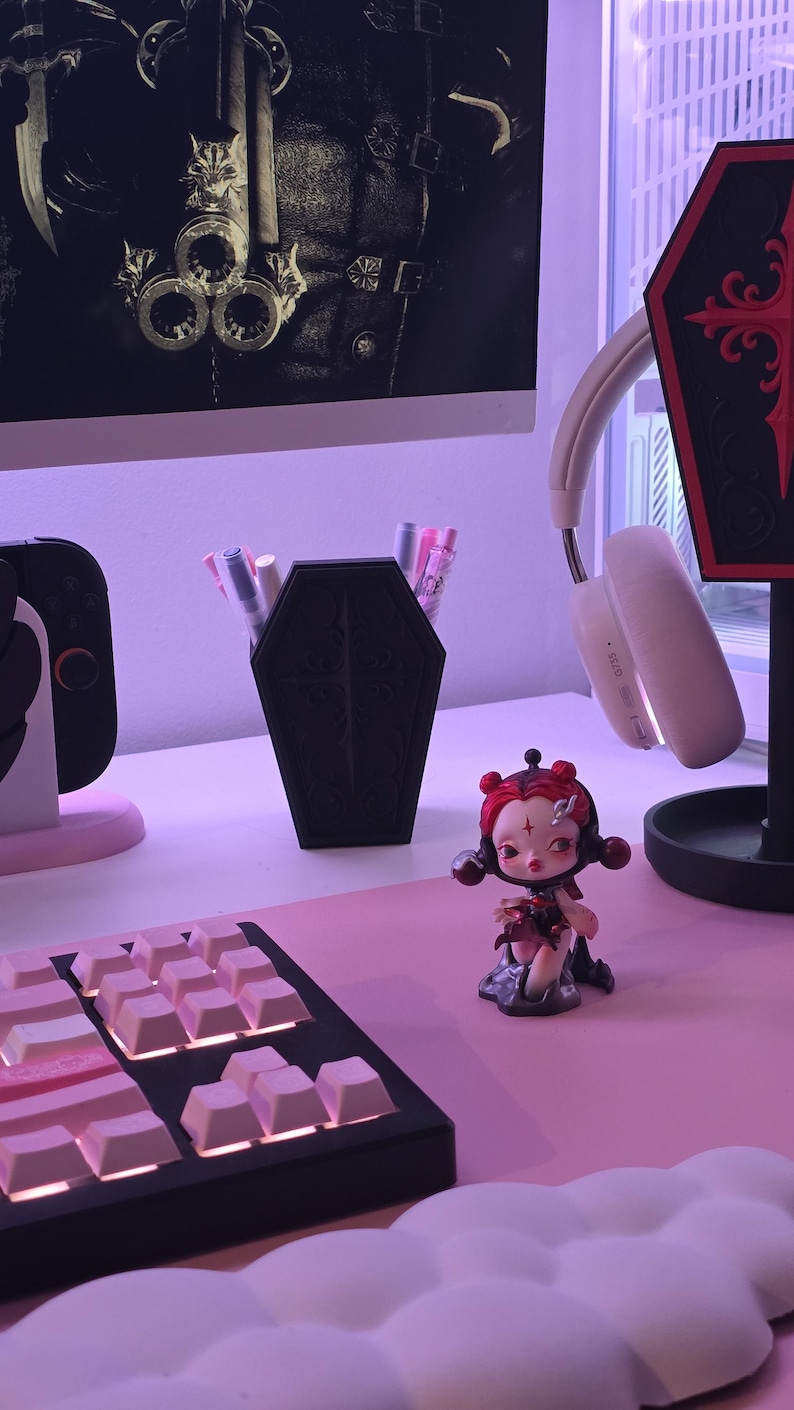 May include: A black coffin-shaped pen holder with a decorative cross design, filled with pens and markers, sits on a pink desk. A small figurine with red hair and a keyboard are also on the desk. A pair of white headphones hangs nearby.