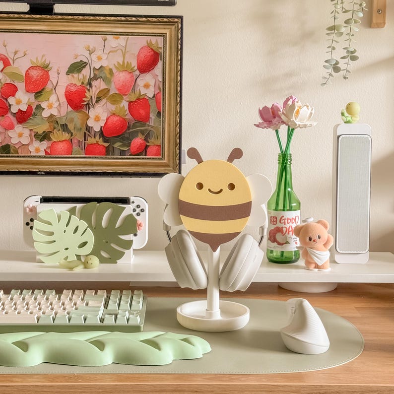 May include: A desk setup featuring a bee-shaped headphone stand with headphones, a keyboard, and a mouse. A framed strawberry painting hangs above a white shelf holding a game console and decorative items. A green desk mat completes the scene.