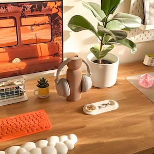 May include: A wooden stand with a pair of grey headphones hanging on it. A small potted succulent plant is in front of the stand. A bright orange keyboard is on a white cloud-shaped wrist rest. A white tray with a gold ring and a silver chain is on a wooden desk.