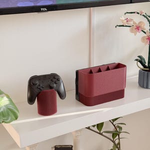 May include: A black video game controller rests on a burgundy cylindrical stand. Beside it, a burgundy organizer with multiple compartments sits on a white shelf. The shelf is against a white wall, with a TV and decorative plants in the background.
