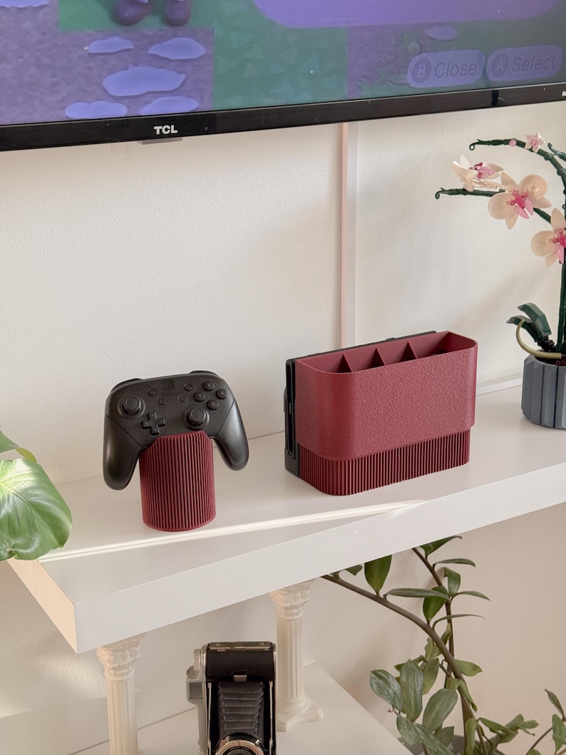 May include: A black video game controller rests on a burgundy cylindrical stand. Beside it, a burgundy organizer with multiple compartments sits on a white shelf. The shelf is against a white wall, with a TV and decorative plants in the background.