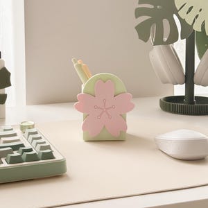 May include: A desk tidy in the shape of a cherry blossom, with a light green base and a pink flower design. It holds pens and pencils. The desk also features a keyboard, mouse, and a headphone stand.