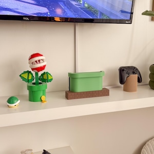 May include: A white shelf with a green and brown video game console stand, a green and red plant-like figure, a brown turtle figurine, a black video game controller, and a brown wooden stand. The shelf is also decorated with two small figurines, one with a pink dress and a crown, and the other with a green dress and a witch's hat.