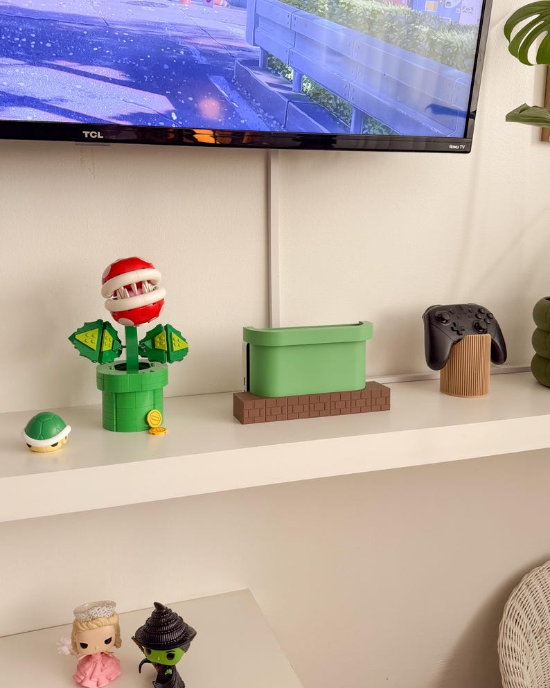 May include: A white shelf with a green and brown video game console stand, a green and red plant-like figure, a brown turtle figurine, a black video game controller, and a brown wooden stand. The shelf is also decorated with two small figurines, one with a pink dress and a crown, and the other with a green dress and a witch's hat.