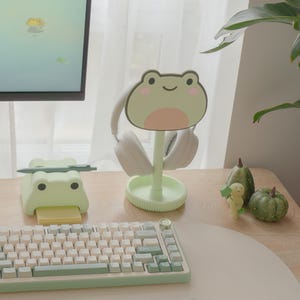 May include: A light green frog-shaped headphone stand with a pair of white headphones. A matching frog-themed sticky note holder and keyboard are also visible. The desk setup includes decorative pumpkins and a small figurine.