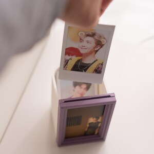Pu&ograve; includere: Una mano tiene in mano una foto polaroid di una persona con i capelli castano chiaro e un gilet giallo. La foto viene inserita in una piccola scatola bianca con una cornice viola. Un'altra foto &egrave; gi&agrave; all'interno della cornice.