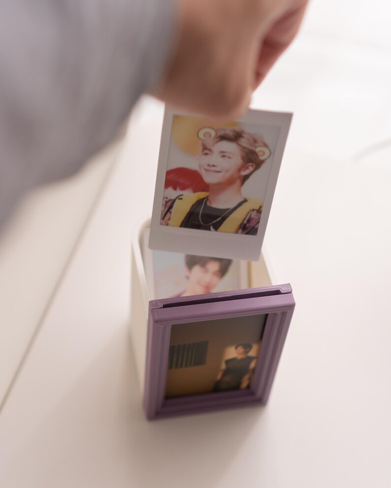 Pu&ograve; includere: Una mano tiene in mano una foto polaroid di una persona con i capelli castano chiaro e un gilet giallo. La foto viene inserita in una piccola scatola bianca con una cornice viola. Un'altra foto &egrave; gi&agrave; all'interno della cornice.