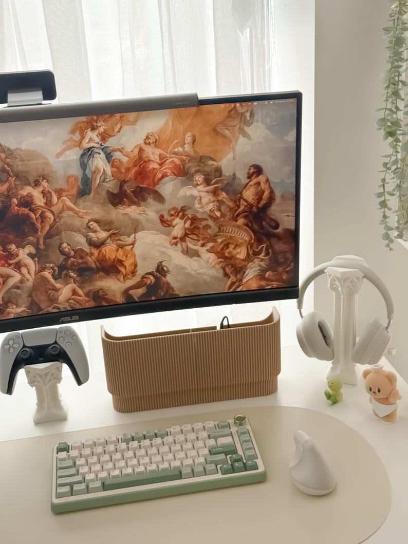 May include: A modern desk setup featuring a computer monitor displaying classical artwork. Includes a white and green keyboard, a white mouse, a beige pencil holder, a white gaming controller, and white headphones. Decorative figurines and a white desk pad complete the scene.