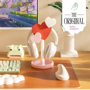 May include: A white headphone stand with a pink base and three heart-shaped decorations. The stand is on a brown desk with a keyboard, mouse, and a small green frog figurine.