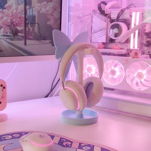 May include: A white and blue butterfly-shaped headphone stand with a pair of white headphones on it. The stand is on a white desk with a pink and blue mousepad and a white mouse.