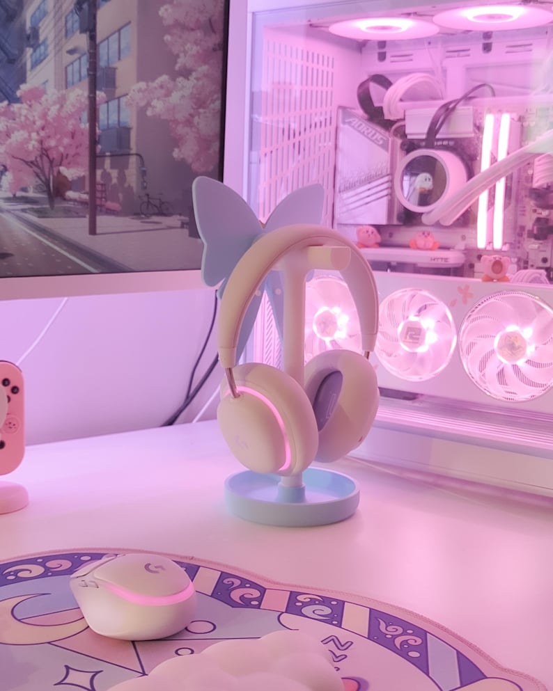 May include: A white and blue butterfly-shaped headphone stand with a pair of white headphones on it. The stand is on a white desk with a pink and blue mousepad and a white mouse.