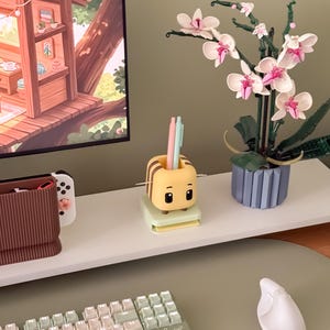 May include: A desk setup featuring a yellow and brown bee-shaped pen holder with pens, a white keyboard, and a white ergonomic mouse. A Lego orchid plant with pink and white flowers sits in a blue pot. A computer monitor displays a treehouse scene.
