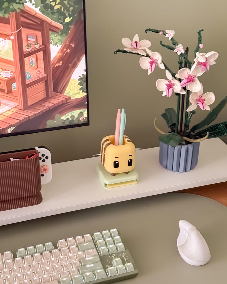 May include: A desk setup featuring a yellow and brown bee-shaped pen holder with pens, a white keyboard, and a white ergonomic mouse. A Lego orchid plant with pink and white flowers sits in a blue pot. A computer monitor displays a treehouse scene.