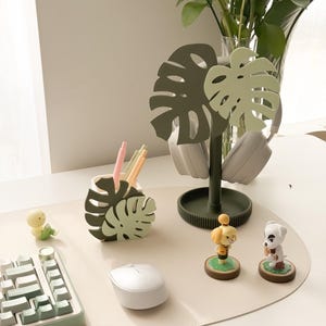 May include: A desk setup featuring a green and white keyboard, a white mouse, and a monstera leaf-shaped pen holder. A headphone stand with monstera leaf design holds a pair of headphones. Two small figurines and a small yellow duck figurine are also present.