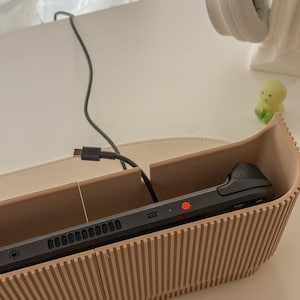 May include: A black handheld gaming console rests inside a beige, ribbed wooden holder. A black charging cable is connected to the console. A pair of white headphones and a small green figurine are visible in the background.