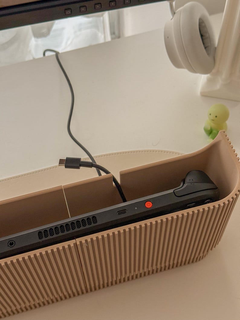 May include: A black handheld gaming console rests inside a beige, ribbed wooden holder. A black charging cable is connected to the console. A pair of white headphones and a small green figurine are visible in the background.