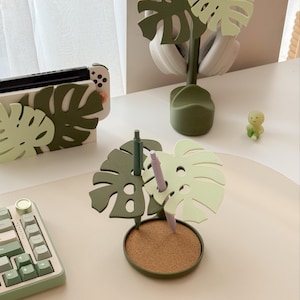 Pu&ograve; includere: Un set di organizzatori da scrivania con un design a foglia di monstera in tonalit&agrave; di verde. Il set include un portapenne con base in sughero, un supporto per cuffie e una custodia per console di gioco portatile. &Egrave; presente anche una piccola statuina verde.